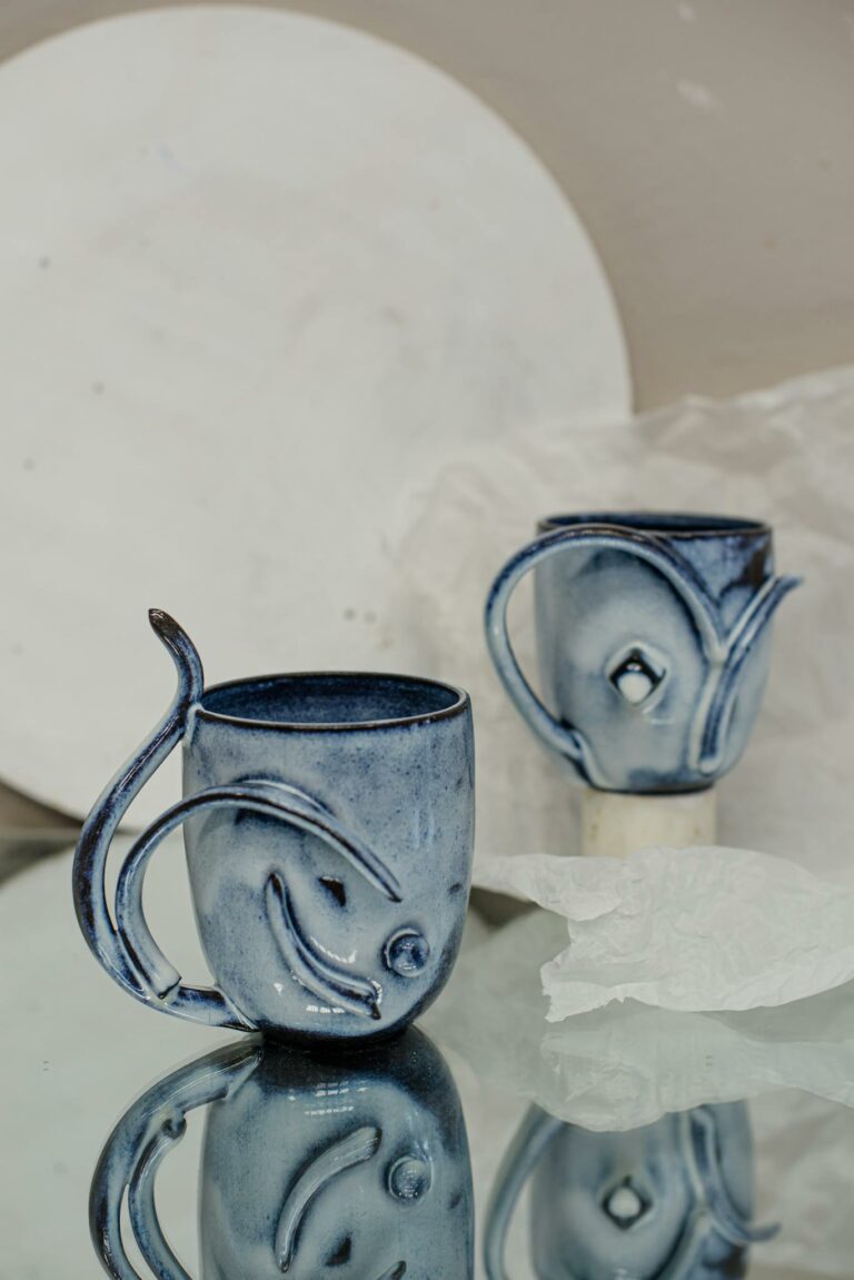 Handcrafted blue ceramic mugs with artistic designs reflecting on glass surface.